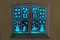 Adventskalenderfenster 23.12.2024 - De Lumina Adventskalenderfenster mit leuchtenden Weihnachtsmotiven wie Sternen, Engeln, Tannenbäumen, einer Krippenszene, römischen Zahlen und dem Schriftzug „DE LUMINA“ in blauem Licht.