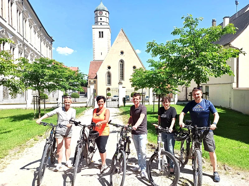 Fünf Personen stehen mit Fahrrädern auf einem Weg vor einer Kirche mit Turm, umgeben von Bäumen und Gebäuden. Im Hintergrund blauer Himmel.