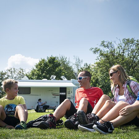 Vier Personen sitzen auf einer Wiese vor Wohnmobilstellplatz in Monheim, im Hintergrund sind Bäume und ein blauer Himmel.