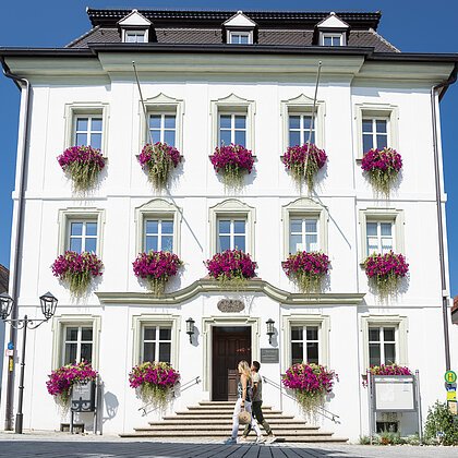 Das Rathaus in Monheim: Ein weißes Gebäude mit mehreren Fenstern, an denen Blumenkästen mit pinken Blumen hängen. Zwei Personen gehen davor vorbei.