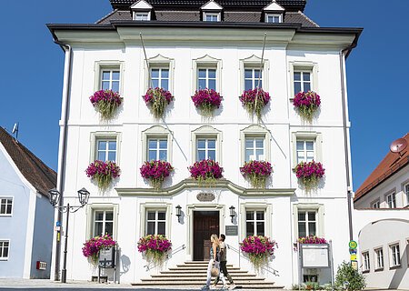 Das Rathaus in Monheim: Ein weißes Gebäude mit mehreren Fenstern, an denen Blumenkästen mit pinken Blumen hängen. Zwei Personen gehen davor vorbei.