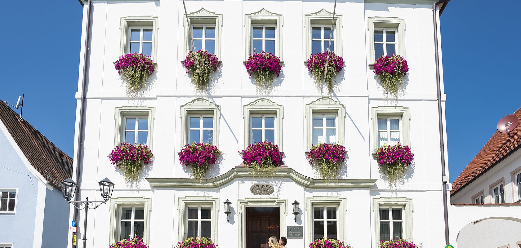 Das Rathaus in Monheim: Ein weißes Gebäude mit mehreren Fenstern, an denen Blumenkästen mit pinken Blumen hängen. Zwei Personen gehen davor vorbei.