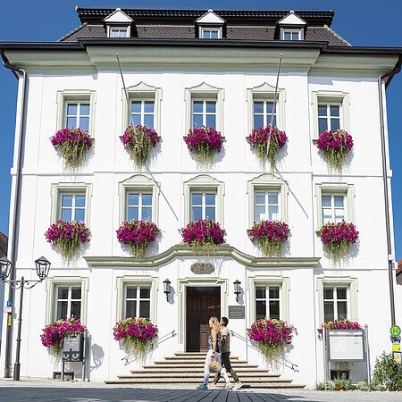 Das Rathaus in Monheim: Ein weißes Gebäude mit mehreren Fenstern, an denen Blumenkästen mit pinken Blumen hängen. Zwei Personen gehen davor vorbei.