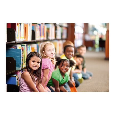 Kinder sitzen in einer Reihe auf dem Boden einer Bibliothek, Bücherregale im Hintergrund.