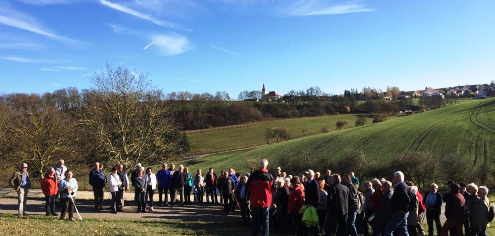 Eine Gruppe von Senioren wandert in der Monheimer Alb. Es stehen Menschen auf einem Weg in einer ländlichen Landschaft unter klarem Himmel.