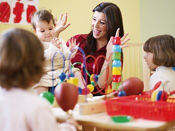 Kinderbetreuung Eine Erwachsene Frau spielt am Tisch mit drei Kindern in einem Raum mit buntem Spielzeug. In der Mitte des Tisches steht ein Motorikwürfel und Bauklötze.