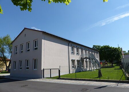 Städtischer Kindergarten in Monheim: ein zweistöckiges Gebäude mit weißer Fassade, Fenstern und Außentreppe, umgeben von einem Zaun und Bäumen. Rechts um das Gebäude ist ein Garten zu sehen. Vorderhalb des Gebäudes verläuft eine Straße.