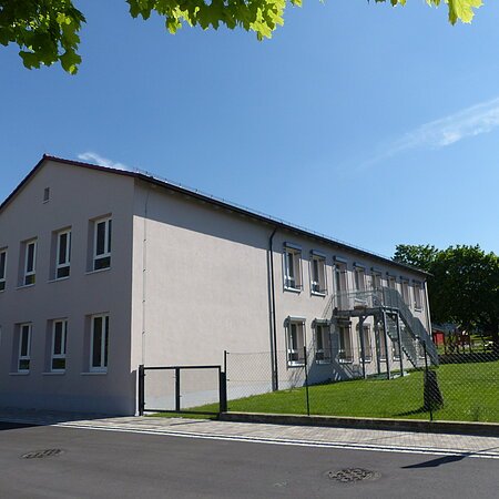 Städtischer Kindergarten in Monheim: ein zweistöckiges Gebäude mit weißer Fassade, Fenstern und Außentreppe, umgeben von einem Zaun und Bäumen. Rechts um das Gebäude ist ein Garten zu sehen. Vorderhalb des Gebäudes verläuft eine Straße.