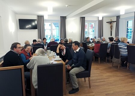 Der Bürgertreff "ZAMSEI" in Monheim: Viele Menschen sitzen bei Kaffee oder Tee in einem Raum an Tischen und unterhalten sich gesellig. Im Hintergrund sind drei Fenster mit Vorhängen und es hängt ein Jesus-Kreuz und ein Fernseher an der Wand.