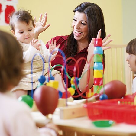 Eine Erwachsene Frau spielt am Tisch mit drei Kindern in einem Raum mit buntem Spielzeug. In der Mitte des Tisches steht ein Motorikwürfel und Bauklötze.