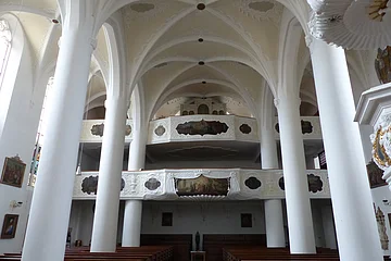 Innenansicht der Stadtpfarrkirche St. Walburga in Monheim, mit hohen weißen Säulen, gewölbter Decke, Emporen mit Verzierungen und Holzbänken.