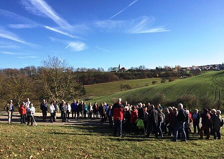 Eine Gruppe von Senioren wandert in der Monheimer Alb. Es stehen Menschen auf einem Weg in einer ländlichen Landschaft unter klarem Himmel.