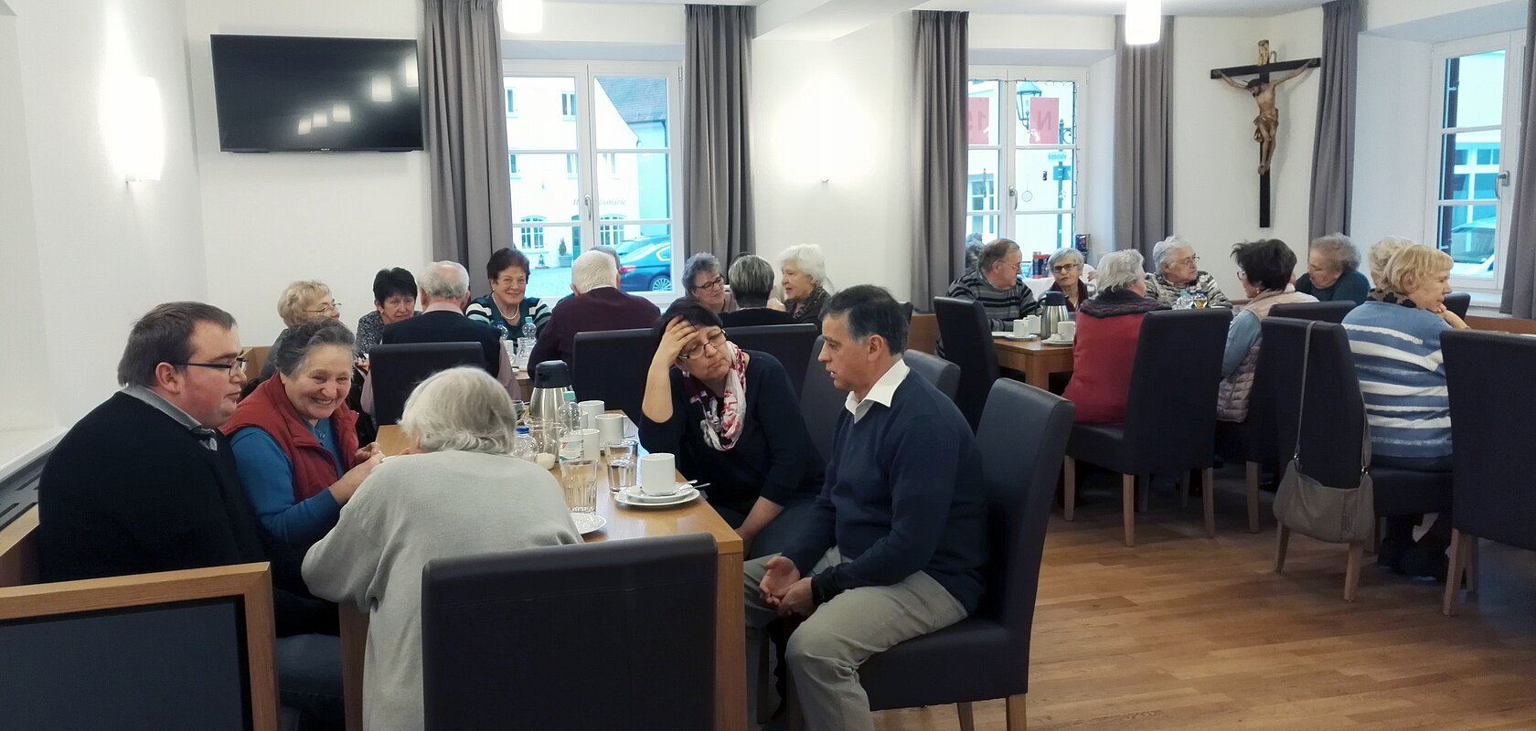 Der Bürgertreff "ZAMSEI" in Monheim: Viele Menschen sitzen bei Kaffee oder Tee in einem Raum an Tischen und unterhalten sich gesellig. Im Hintergrund sind drei Fenster mit Vorhängen und es hängt ein Jesus-Kreuz und ein Fernseher an der Wand.