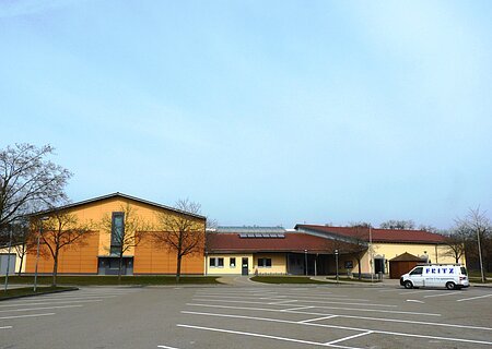 Stadt-/Mehrzweckhalle in Monheim: Ein großes, modernes Gebäude mit gelber Fassade und rotem Dach und mehreren Bäumen. Vorderhalb des Gebäudes ist ein großer Parkplatz, auf dem ein weißes Auto steht.