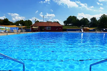 Das Freibad in Monheim: Schwimmbecken mit klarem Wasser, umgeben von Liegestühlen, einem roten Gebäude und Umkleidekabinen im Hintergrund.