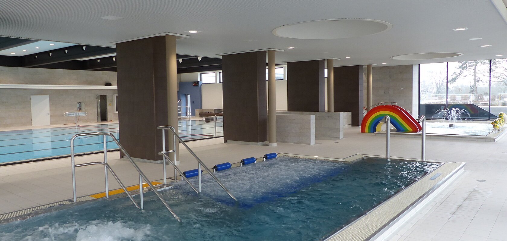 Innenansicht eines Schwimmbads mit mehreren Becken, Rutsche in Regenbogenfarben und Wasserspielen.