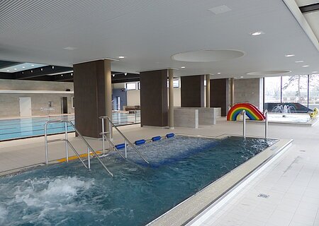 Innenansicht eines Schwimmbads mit mehreren Becken, Rutsche in Regenbogenfarben und Wasserspielen.