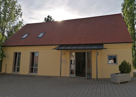 Das Haus Martin Luther in Monheim, ein evangelisches Gemeindehaus: Ein gelbes Gebäude mit rotem Satteldach, zwei Fenstern und einer offenen Eingangstür, umgeben von Bäumen. Vor dem Haus steht ein bepflanztes Beet.