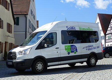 Weißer Kleinbus steht auf einem gepflasterten Platz, in der Monheimer Innenstadt . Auf dem Bus ist die Aufschrift "SoMit BürgerBus" und "wir verbinden die Monheimer Alb".