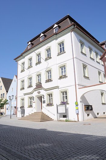 Das Rathaus der Stadt Monheim, am Marktplatz. Ein weißes, dreistöckiges Gebäude mit Satteldach und mehreren Fenstern, im Vordergrund gepflasterter Marktplatz.
