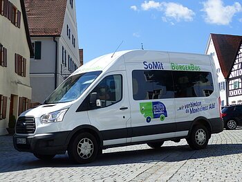 SoMit Bürgerbus Weißer Kleinbus steht auf einem gepflasterten Platz, in der Monheimer Innenstadt . Auf dem Bus ist die Aufschrift "SoMit BürgerBus" und "wir verbinden die Monheimer Alb".
