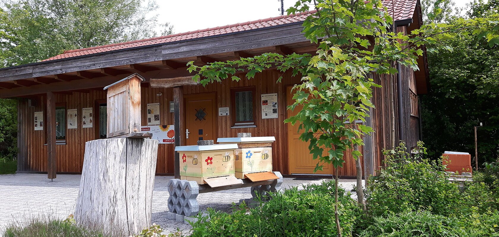 Lehrbienenstand in Monheim: ein Holzgebäude mit rotem Dach, umgeben von Bäumen und Sträuchern, im Vordergrund ein Baumstumpf und Bienenkästen.
