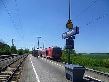 Der Bahnhof Otting-Weilheim mit rotem Zug auf dem Gleis, blauer Himmel, grüne Landschaft im Hintergrund.