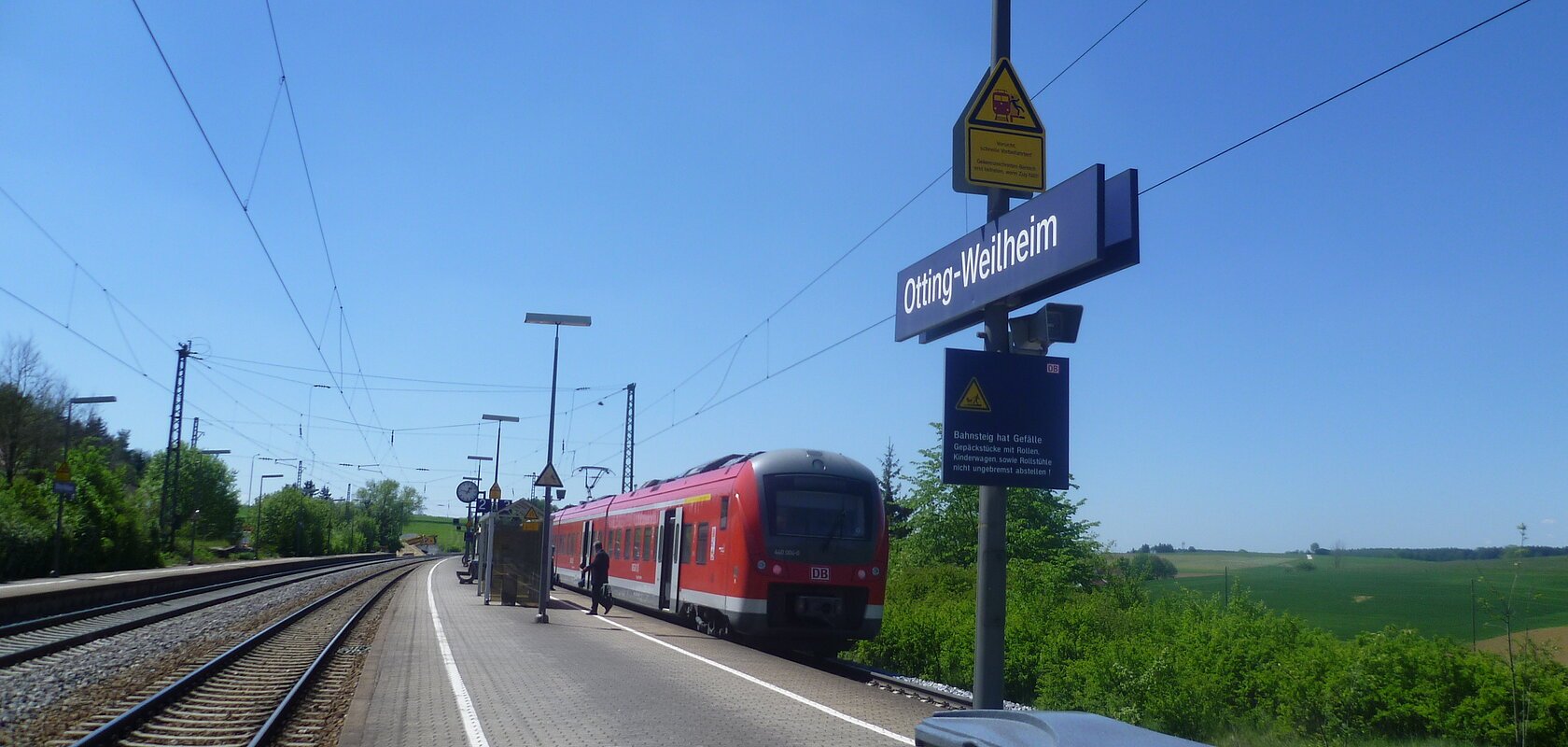 Der Bahnhof Otting-Weilheim mit rotem Zug auf dem Gleis, blauer Himmel, grüne Landschaft im Hintergrund.