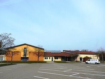 Stadt-/Mehrzweckhalle Monheim Stadt-/Mehrzweckhalle in Monheim: Ein großes, modernes Gebäude mit gelber Fassade und rotem Dach und mehreren Bäumen. Vorderhalb des Gebäudes ist ein großer Parkplatz, auf dem ein weißes Auto steht.