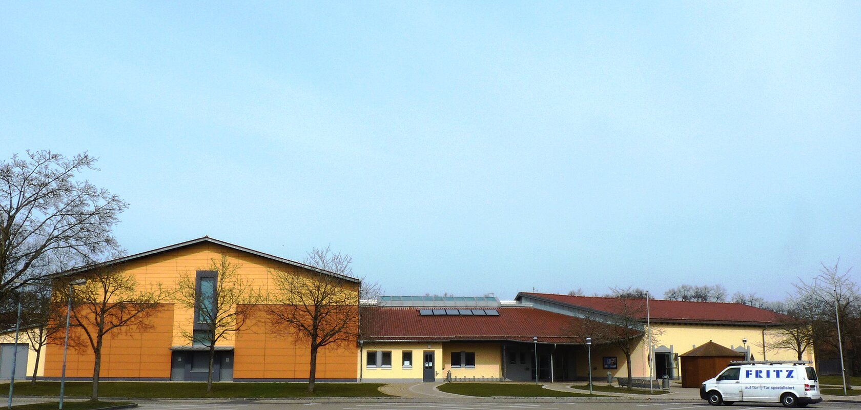 Stadt-/Mehrzweckhalle in Monheim: Ein großes, modernes Gebäude mit gelber Fassade und rotem Dach und mehreren Bäumen. Vorderhalb des Gebäudes ist ein großer Parkplatz, auf dem ein weißes Auto steht.