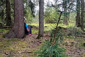 Person sitzt im Wald auf dem Boden, lehnt an einem Baum. Umgebung aus Bäumen und Moos.
