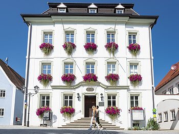 Das Rathaus der Drei-Stämme-Stadt Monheim Das Rathaus in Monheim: Ein weißes dreistöckiges Gebäude mit mehreren Fenstern, an denen Blumenkästen mit pinken Blumen hängen. Zwei Personen gehen davor vorbei.