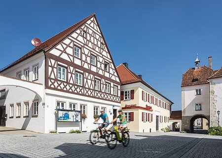 Tourist-Information der Stadt Monheim/Monheimer Alb: ein Fachwerkhaus mit drei Stockwerken, weißen Wänden und braunen Balken. Davor steht ein Schaukasten und zwei Radfahrer fahren auf gepflasterter Straße vorbei. Rechts davon ein gelbes zweistöckiges Gebäude mit roten Fensterläden.