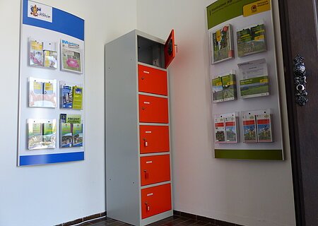 Ein orange-grauer Indoor-Ladeschrank für eBike-Akkus mit insgesamt sechs Fächern und einem offenen Fach steht zwischen zwei Wanddisplays mit Broschüren in dem Vorraum der Touristinformation Monheim.