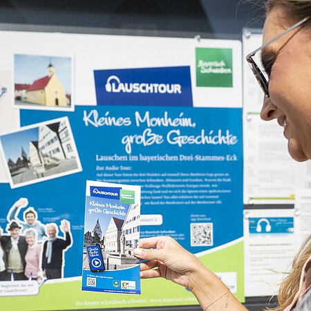 Eine Person steht vor der Touristinformation Monheim vor einer Informationstafel zur Lauschtour in Monheim und betrachtet die dazugehörige Broschüre, die sie in der Hand hält.