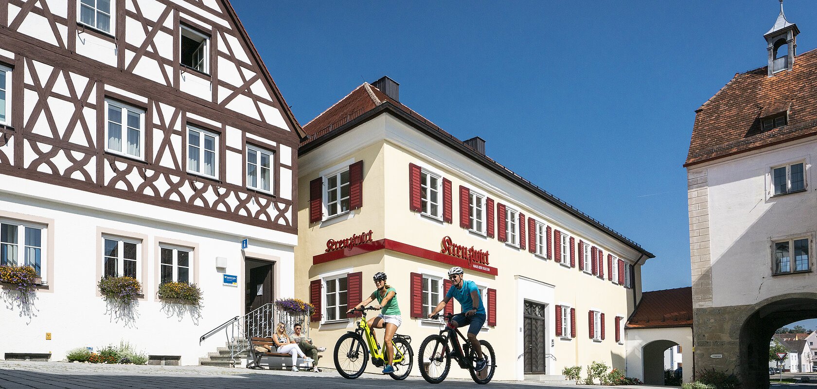 Links im Bild Tourist-Information der Stadt Monheim/Monheimer Alb: ein Fachwerkhaus mit drei Stockwerken, weißen Wänden und braunen Balken. Rechts davon ein gelbes zweistöckiges Gebäude mit roten Fensterläden. Auf einer Sitzbank sitzen zwei Personen. Davor fahren zwei Radfahrer auf gepflasterter Straße vorbei.