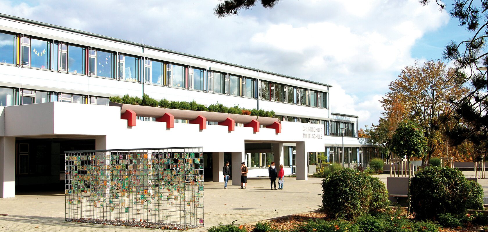 Schulgebäude der Grund- und Mittelschule Monheim, mit Flachdach, Glasfassade und Eingangsbereich, umgeben von Bäumen und Sträuchern. Auf dem Hof stehen vier Personen bei klarem Himmel.