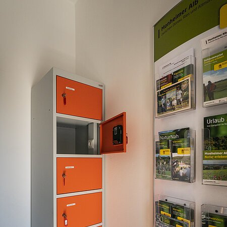 Ein orange-grauer Indoor-Ladeschrank für eBike-Akkus mit insgesamt sechs Fächern und einem offenen Fach steht neben einem Wanddisplay mit Broschüren in dem Vorraum der Touristinformation Monheim.
