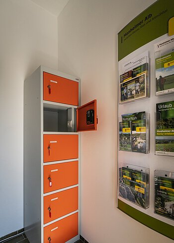 Ein orange-grauer Indoor-Ladeschrank für eBike-Akkus mit insgesamt sechs Fächern und einem offenen Fach steht neben einem Wanddisplay mit Broschüren in dem Vorraum der Touristinformation Monheim.