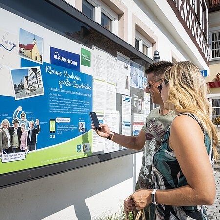 Zwei Personen vor einem weißen Gebäude, der Touristinformation Monheim betrachten eine Informationstafel mit einer Karte und Texten zur Lauschtour in Monheim und haben ein Smartphone in der Hand, mit dem sie einen Code abscannen.