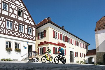 Links im Bild Tourist-Information der Stadt Monheim/Monheimer Alb: ein Fachwerkhaus mit drei Stockwerken, weißen Wänden und braunen Balken. Rechts davon ein gelbes zweistöckiges Gebäude mit roten Fensterläden. Auf einer Sitzbank sitzen zwei Personen. Davor fahren zwei Radfahrer auf gepflasterter Straße vorbei.