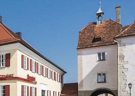 Tourist-Information der Stadt Monheim/Monheimer Alb: ein Fachwerkhaus mit drei Stockwerken, weißen Wänden und braunen Balken. Davor steht ein Schaukasten und zwei Radfahrer fahren auf gepflasterter Straße vorbei. Rechts davon ein gelbes zweistöckiges Gebäude mit roten Fensterläden.
