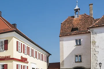 Tourist-Information der Stadt Monheim/Monheimer Alb: ein Fachwerkhaus mit drei Stockwerken, weißen Wänden und braunen Balken. Davor steht ein Schaukasten und zwei Radfahrer fahren auf gepflasterter Straße vorbei. Rechts davon ein gelbes zweistöckiges Gebäude mit roten Fensterläden.