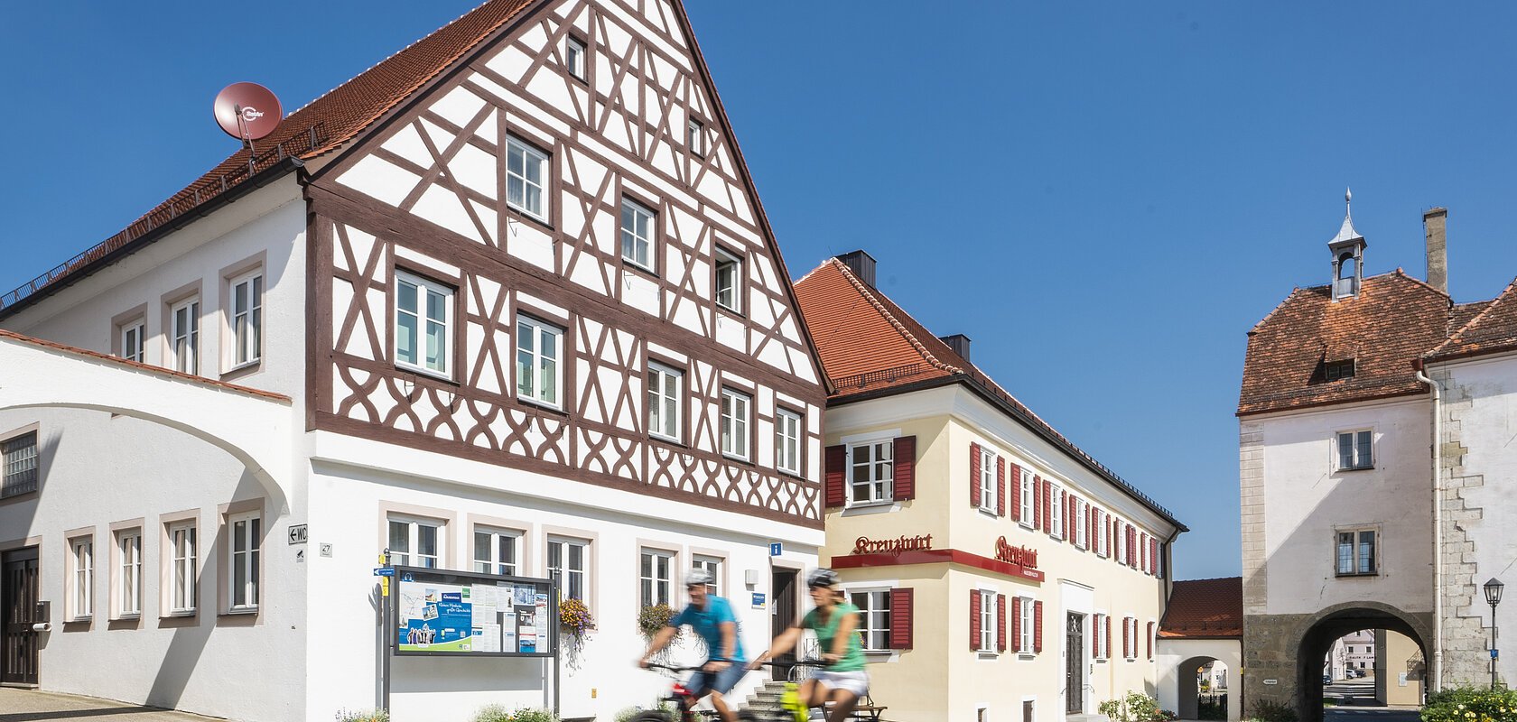 Tourist-Information der Stadt Monheim/Monheimer Alb: ein Fachwerkhaus mit drei Stockwerken, weißen Wänden und braunen Balken. Davor steht ein Schaukasten und zwei Radfahrer fahren auf gepflasterter Straße vorbei. Rechts davon ein gelbes zweistöckiges Gebäude mit roten Fensterläden.