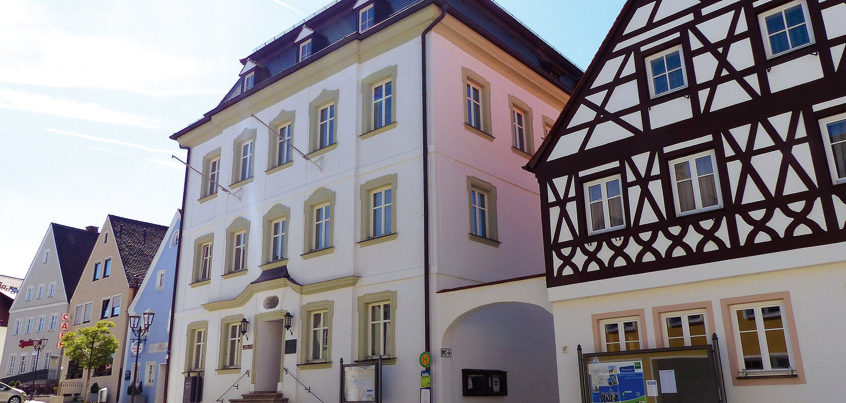 Straßenansicht der Monheimer Innenstadt: Im Vordergrund das Rathaus der Stadt Monheim, mit weiteren mehreren Gebäuden, darunter ein Fachwerkhaus, das man Schindlerhaus nennt.