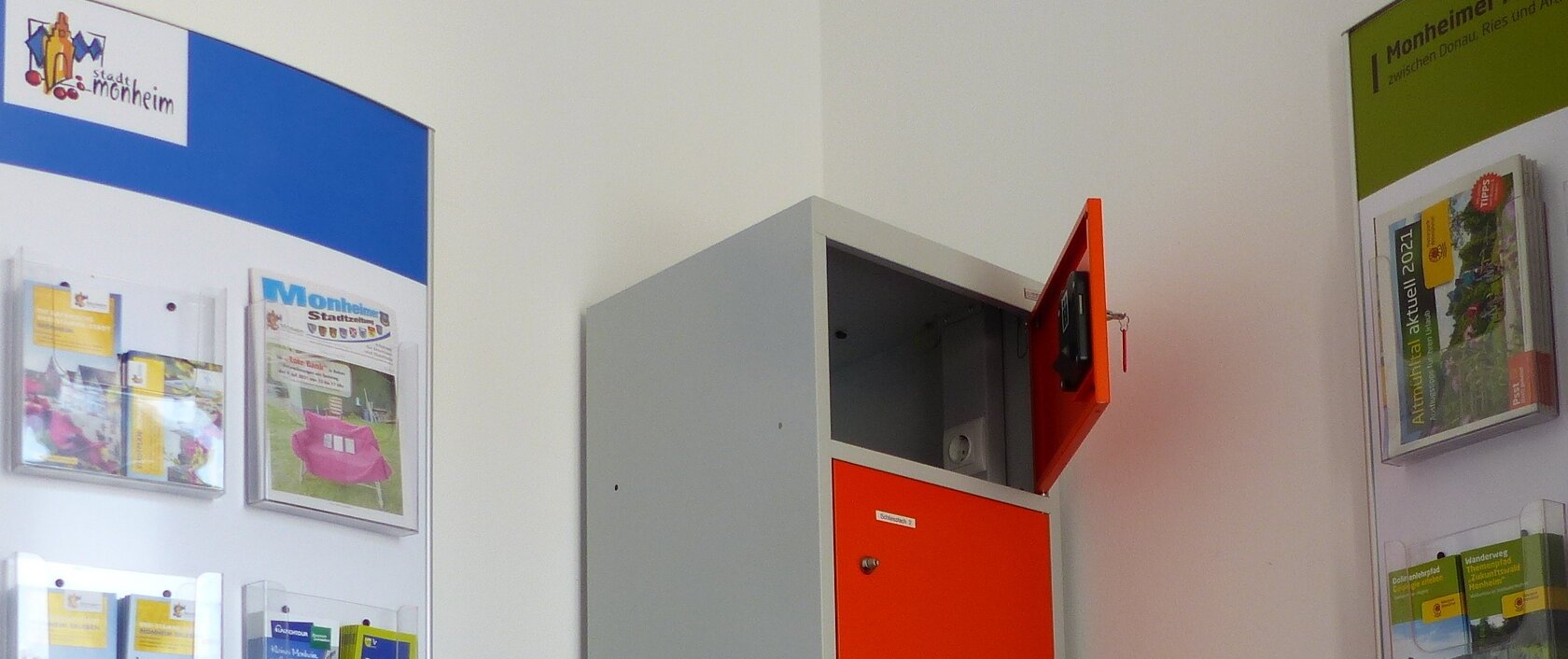 Ein orange-grauer Indoor-Ladeschrank für eBike-Akkus mit insgesamt sechs Fächern und einem offenen Fach steht zwischen zwei Wanddisplays mit Broschüren in dem Vorraum der Touristinformation Monheim.
