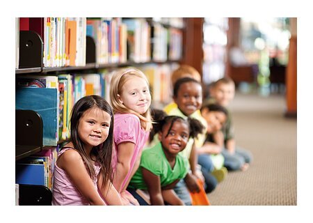 Kinder sitzen in einer Reihe auf dem Boden einer Bibliothek, Bücherregale im Hintergrund.