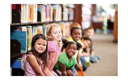 Kinder sitzen in einer Reihe auf dem Boden einer Bibliothek, Bücherregale im Hintergrund.