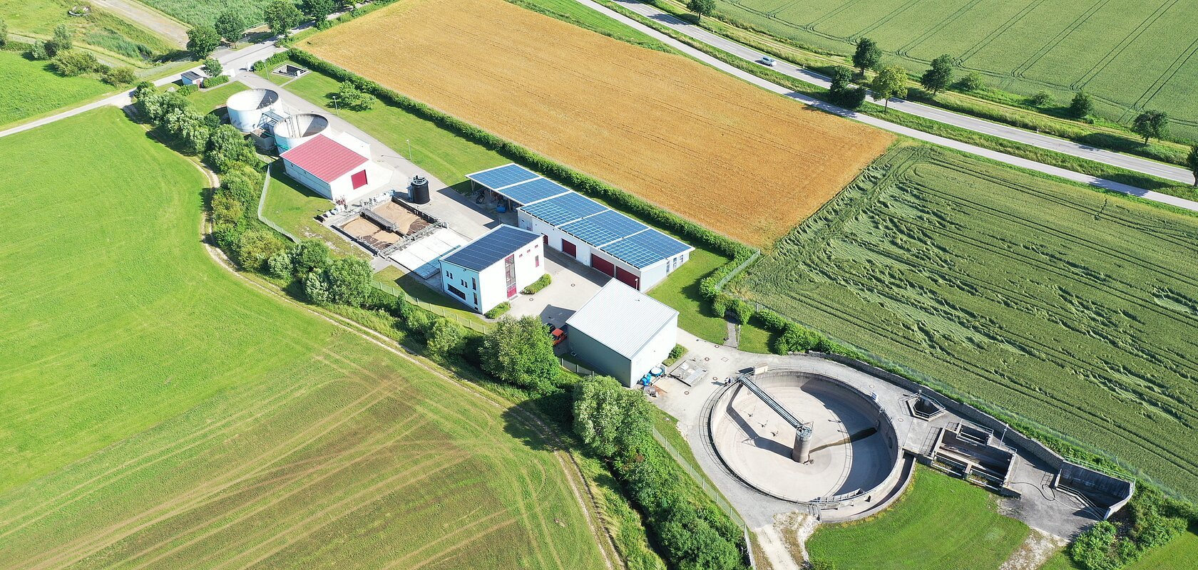 Luftaufnahme der städtische Kläranlage Monheim mit Solarpaneelen auf den Dächern, umgeben von grünen Feldern und Straßen.