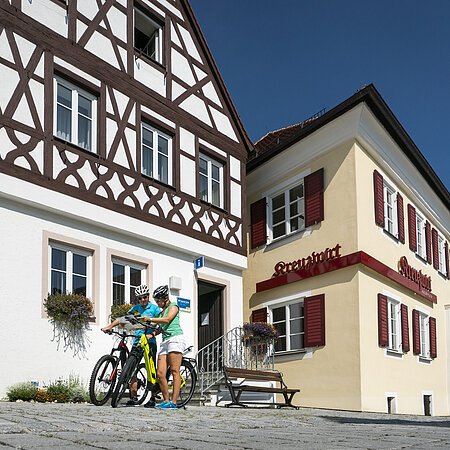 Tourist-Information der Stadt Monheim/Monheimer Alb: ein Fachwerkhaus mit drei Stockwerken, weißen Wänden und braunen Balken. Rechts davon ein gelbes zweistöckiges Gebäude mit roten Fensterläden. Zwei Personen mit Fahrrädern stehen davor und schauen eine Broschüre an.